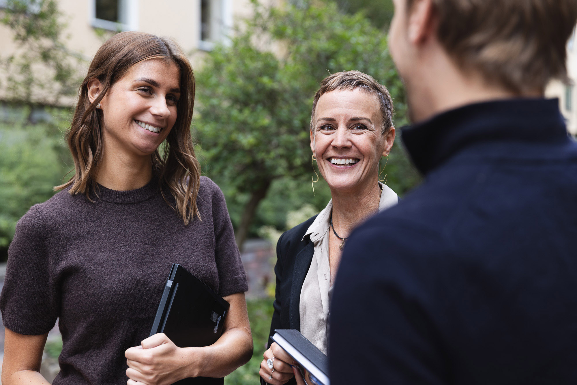 Two women smiling and talking outdoors while holding books and a tablet, with a third person seen from behind.