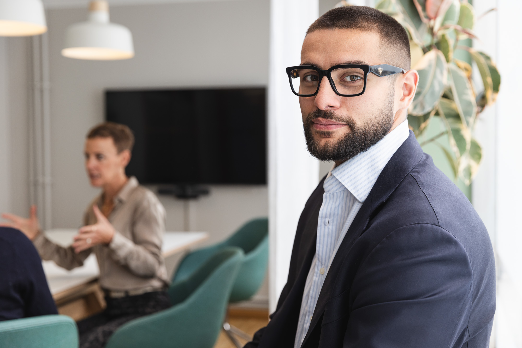 Man with glasses and beard wearing a navy blazer looking at the camera in a modern office with a blurred woman talking in the background.