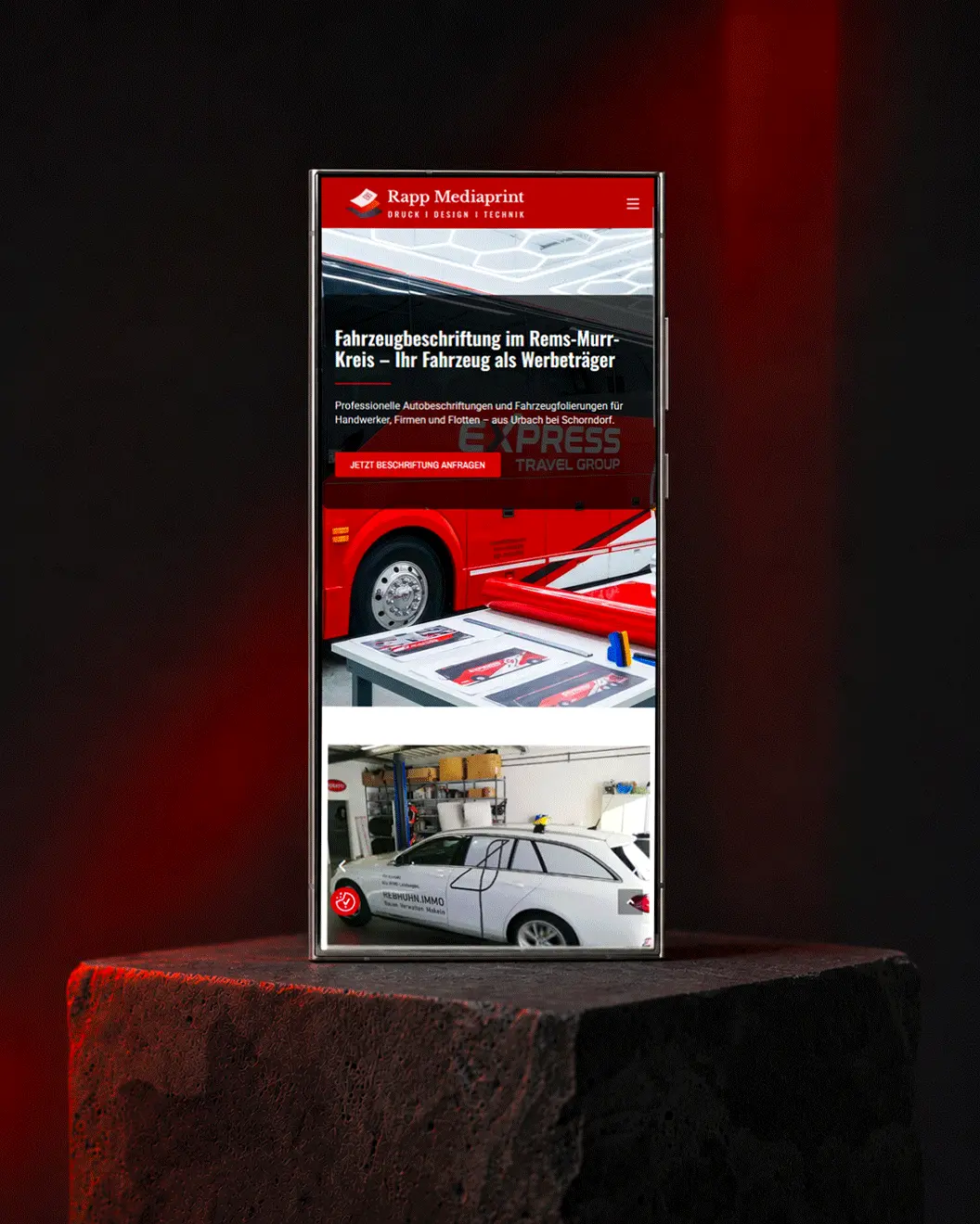 Smartphone-Mockup der Webdesign-Projektseite von Rapp Mediaprint aus Urbach bei Schorndorf für Druck, Fahrzeugbeschriftung und Werbetechnik auf dunklem Steinpodest mit rotem Lichtakzent, umgesetzt von dencraft.