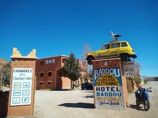 Eingangsschild eines Hotels mit einer gelben Autoskulptur und einer Möwe darauf, ein Motorrad ist daneben geparkt, im Hintergrund ein Gebäude und ein klarer blauer Himmel.