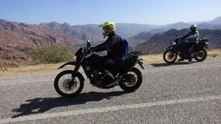 Zwei Motorradfahrer auf einer Straße vor einer gebirgigen Wüstenlandschaft bei klarem Himmel.