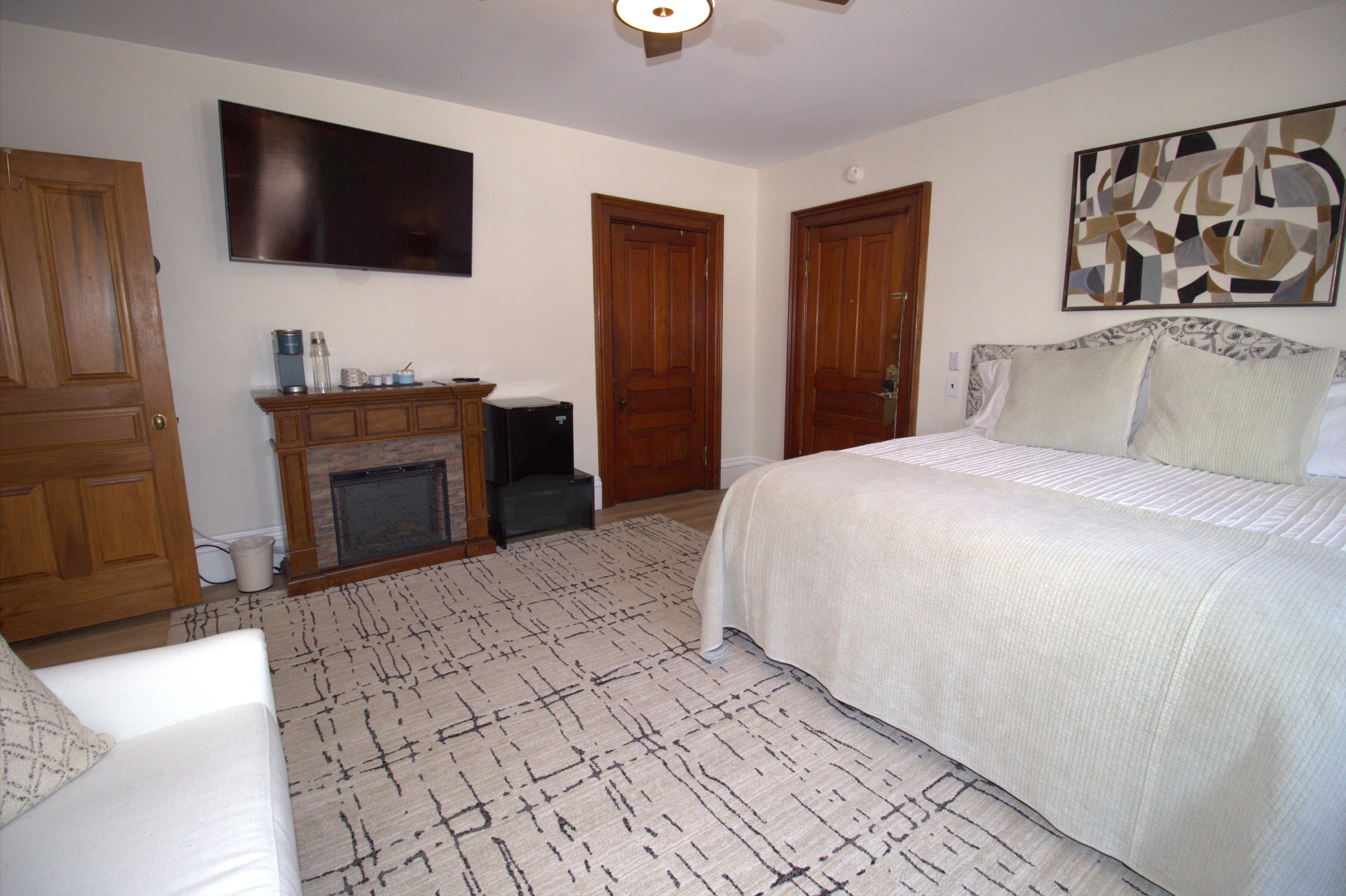 Bedroom with a large bed covered in beige bedding, patterned area rug, wooden doors, fireplace mantel with coffee maker, and a mounted flat screen TV.