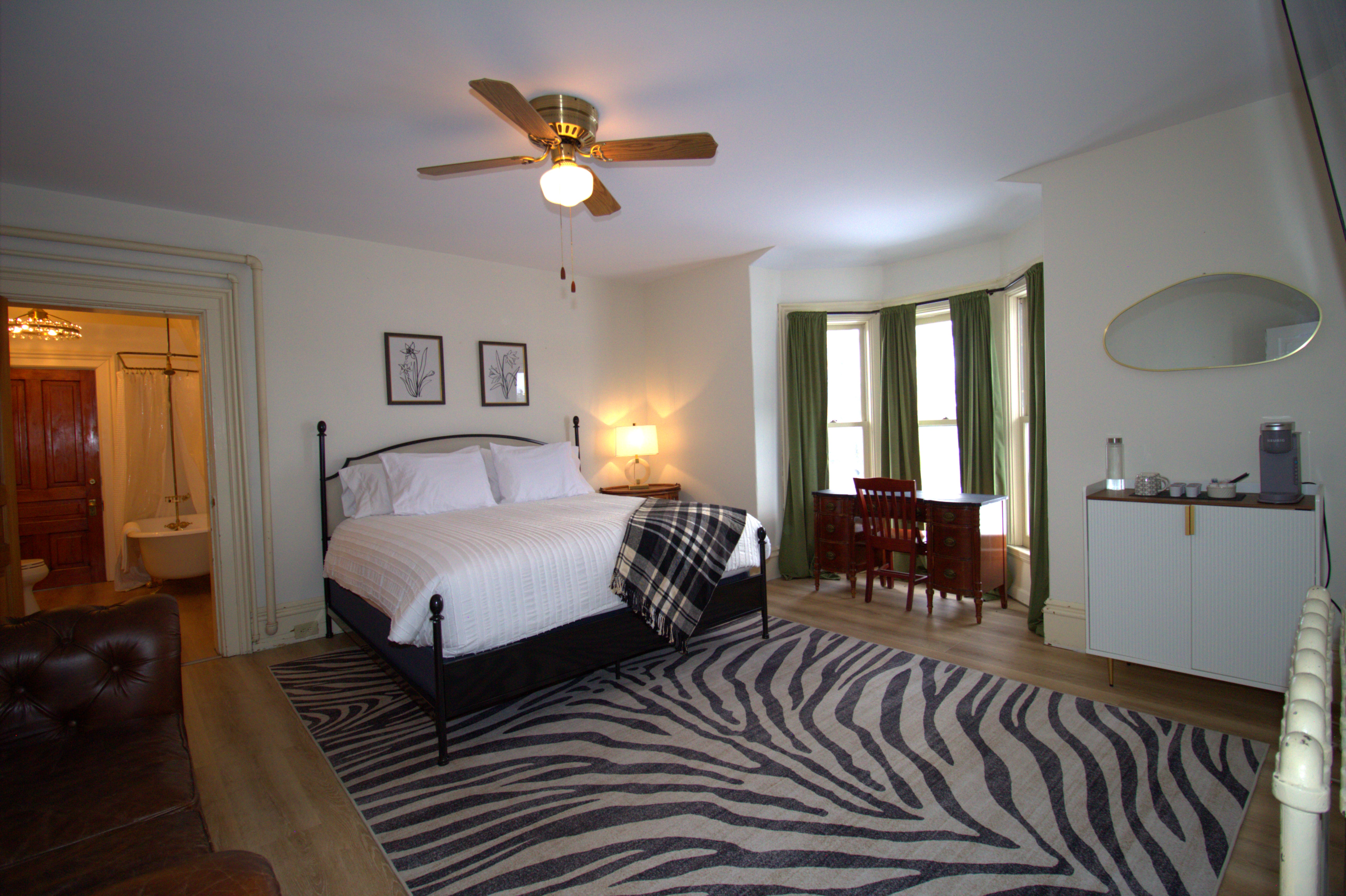Spacious bedroom with a black metal bed, white bedding, a plaid throw, wooden desk by bay windows with green curtains, a leather sofa, zebra-print rug, and an adjacent bathroom visible.
