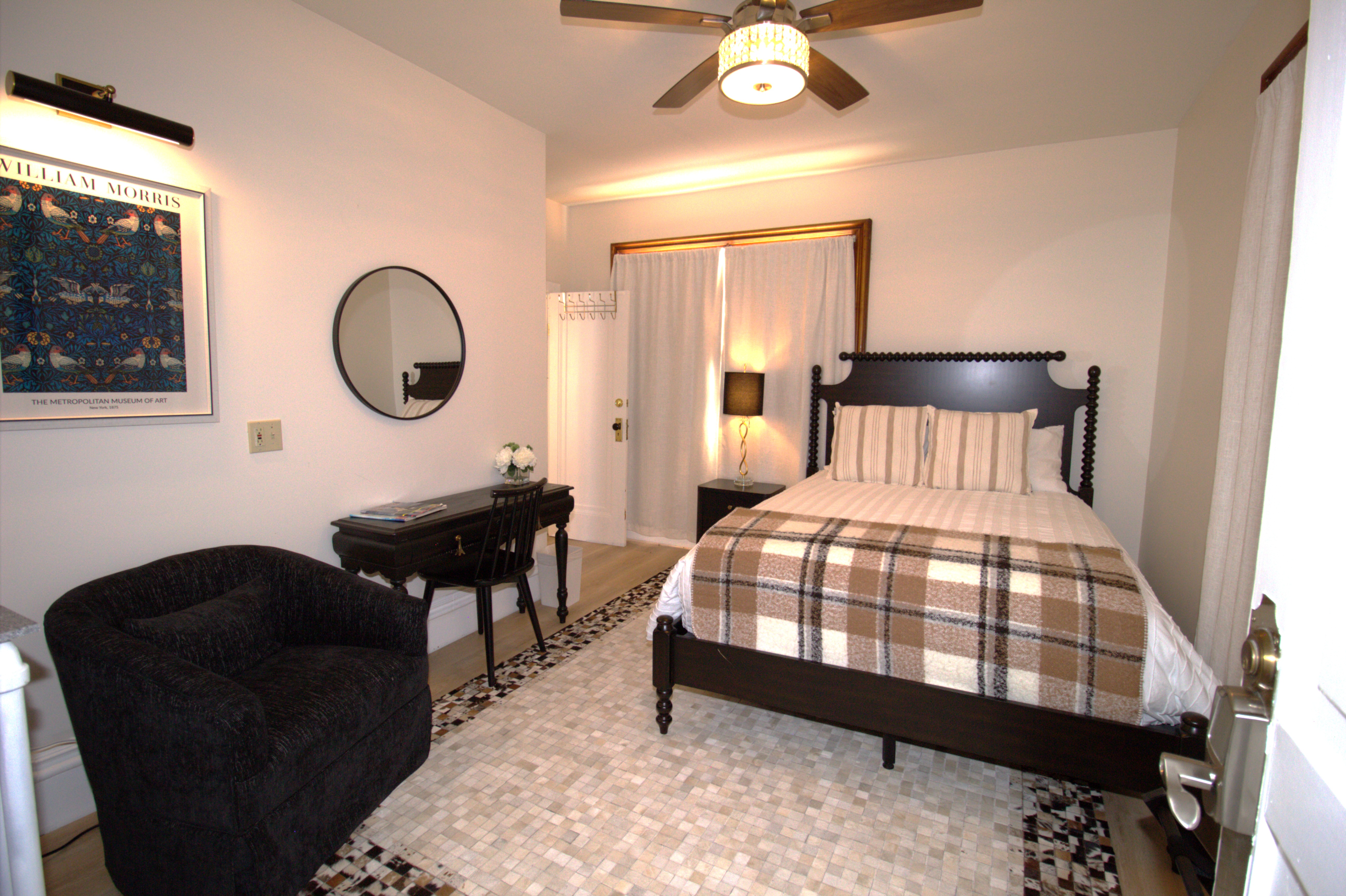 Cozy bedroom with dark wood bed frame, plaid blanket, black armchair, small desk with chair, round wall mirror, and a lamp on a nightstand.