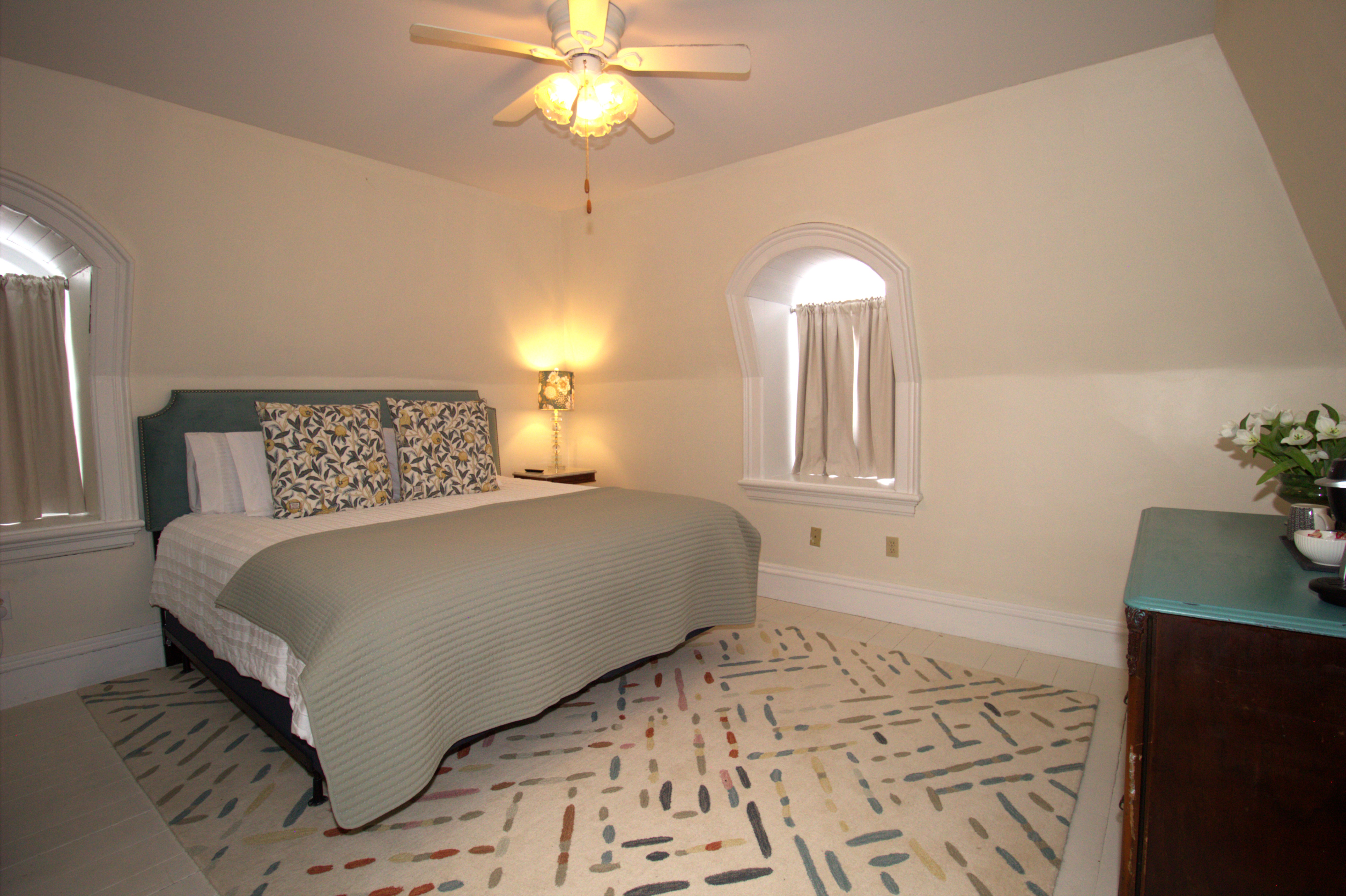 Cozy bedroom with a large bed featuring floral pillows, a green quilt, two arched windows with curtains, a bedside lamp, and a patterned rug.