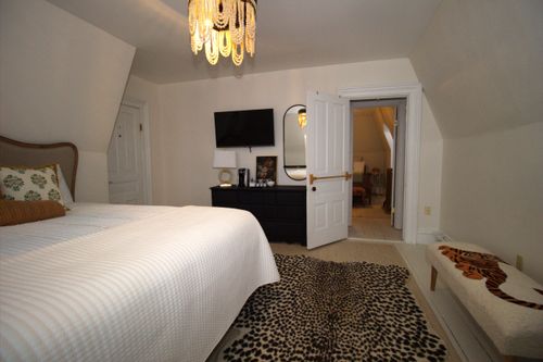 Cozy bedroom with white quilted bedspread, leopard print rug, black dresser, wall-mounted TV, and beaded chandelier.