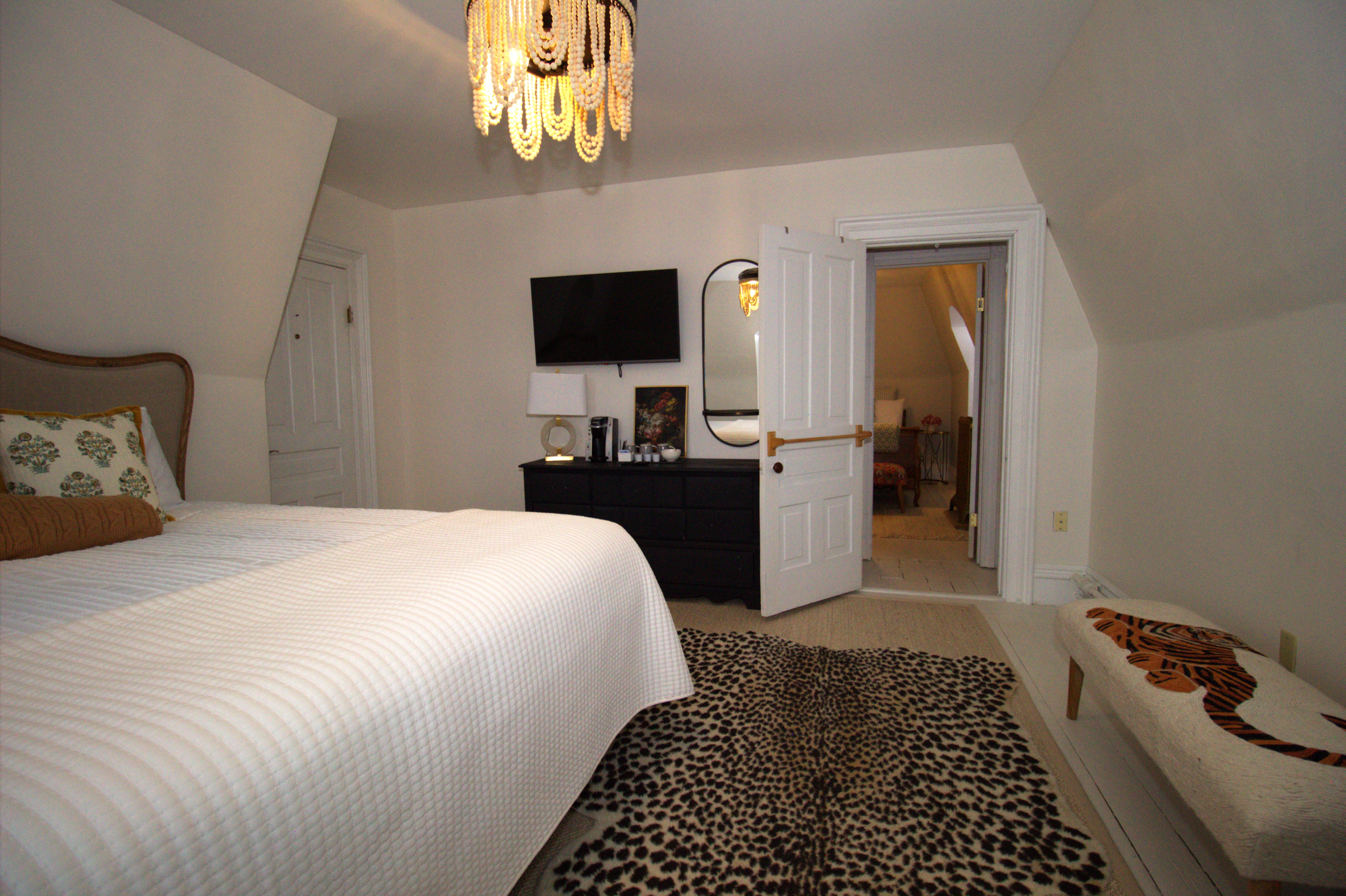 Cozy bedroom with white quilted bedspread, leopard print rug, black dresser, wall-mounted TV, and beaded chandelier.
