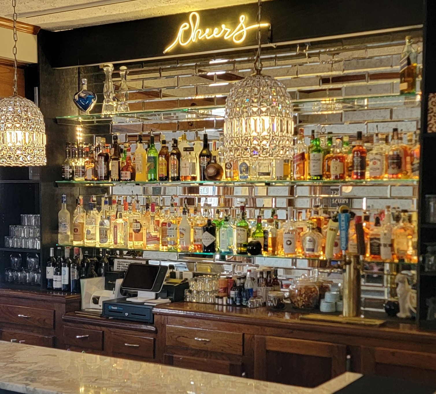 Bar counter with wooden drawers, illuminated liquor bottles on glass shelves, two crystal pendant lights, and a neon 'cheers' sign on a mirrored wall.
