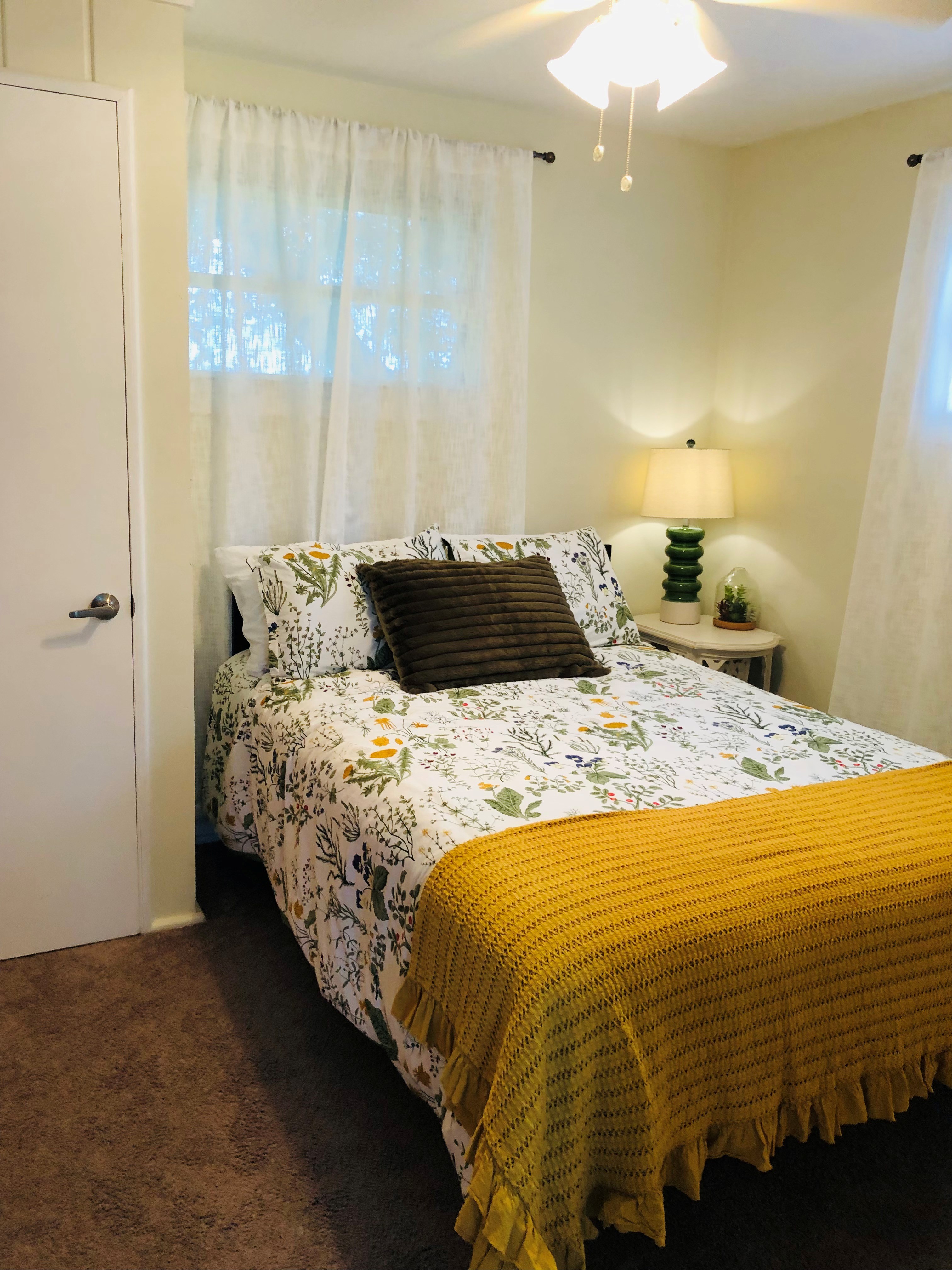 Cozy bedroom with a white bed, decorative pillows including one that reads 'To the Beach', a small white nightstand with a lamp, and a red patterned ottoman.