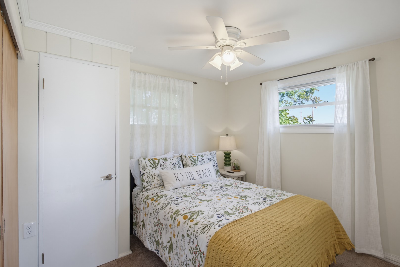 Cozy bedroom with a white bed, decorative pillows including one that reads 'To the Beach', a small white nightstand with a lamp, and a red patterned ottoman.