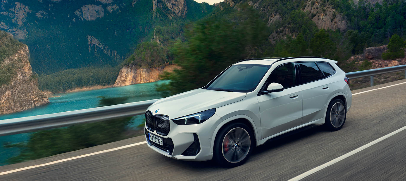 Weißer BMW iX1 fährt dynamisch auf kurviger Küstenstraße entlang türkisblauer Felslandschaft – BMW Wolter & Steiner in Neubrandenburg, Waren und Prenzlau