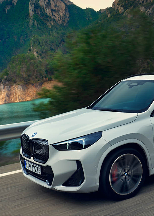 Weißer BMW iX1 fährt dynamisch auf kurviger Küstenstraße entlang türkisblauer Felslandschaft – BMW Wolter & Steiner in Neubrandenburg, Waren und Prenzlau