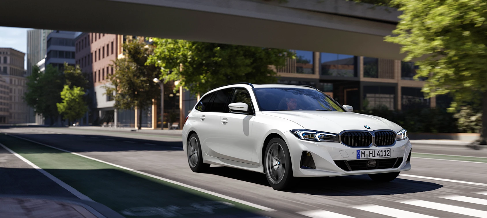 Weißer BMW 3er Touring fährt dynamisch durch urbane Straße mit moderner Architektur – BMW Wolter & Steiner in Neubrandenburg, Waren und Prenzlau