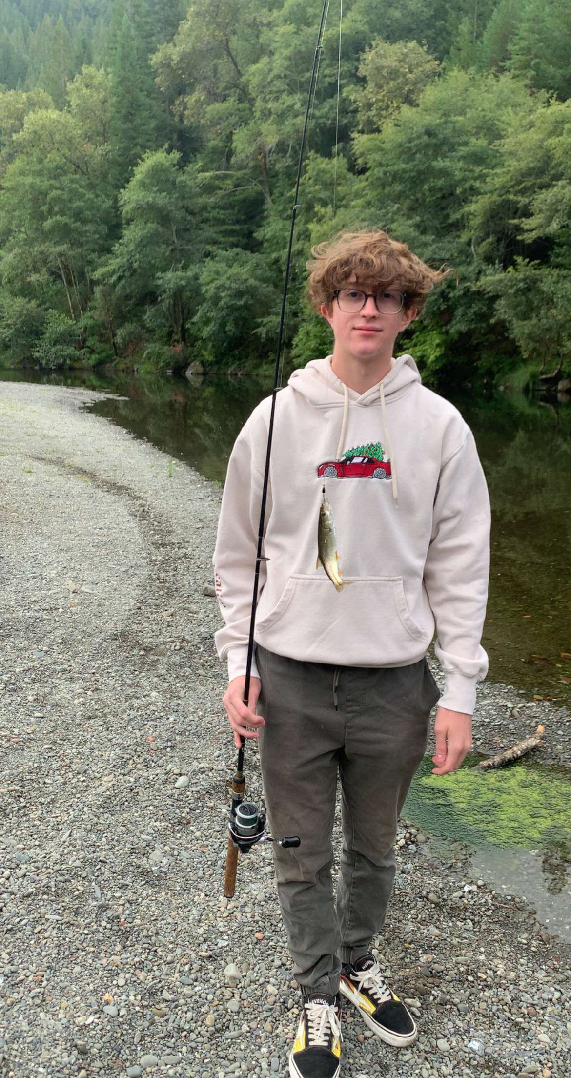 Young man with curly hair and glasses standing on a gravel riverbank holding a fishing rod with a small fish caught on the line, surrounded by green trees.