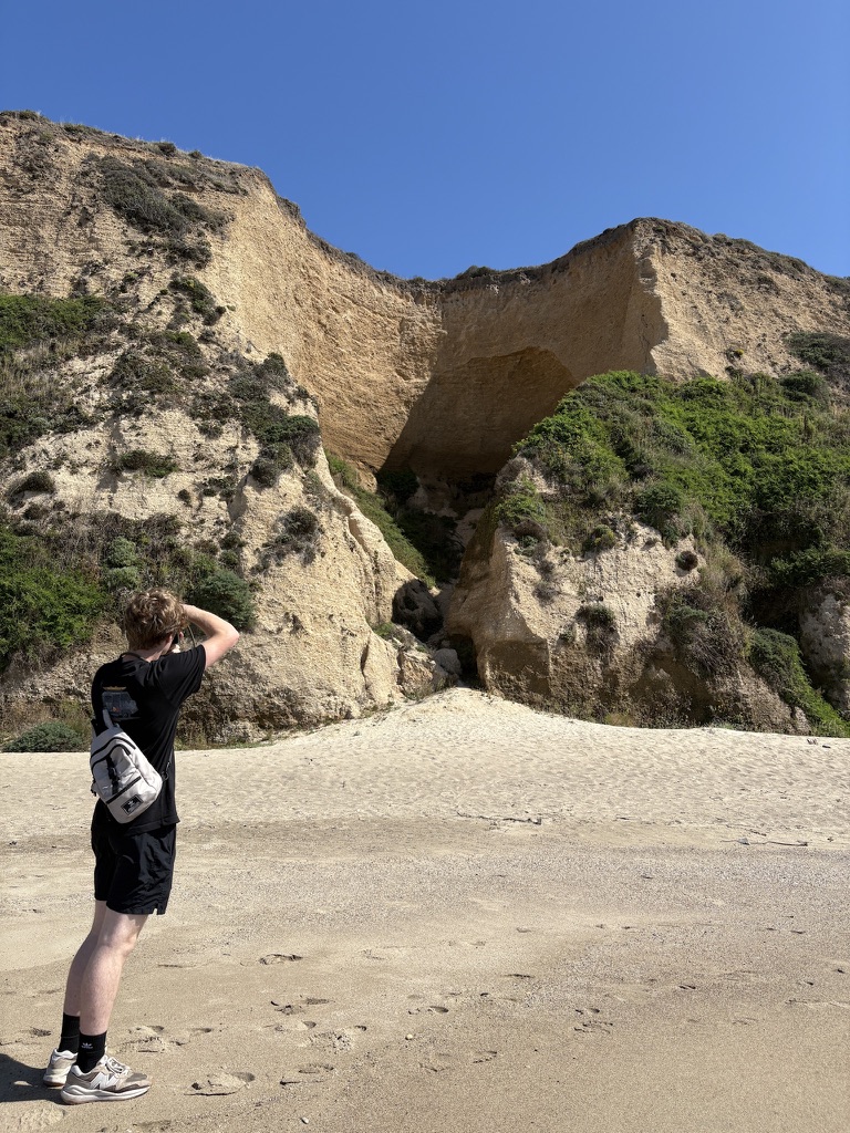 Person wearing black outfit and light backpack photographing a large sandy cliff with green vegetation under clear blue sky on a sandy beach.