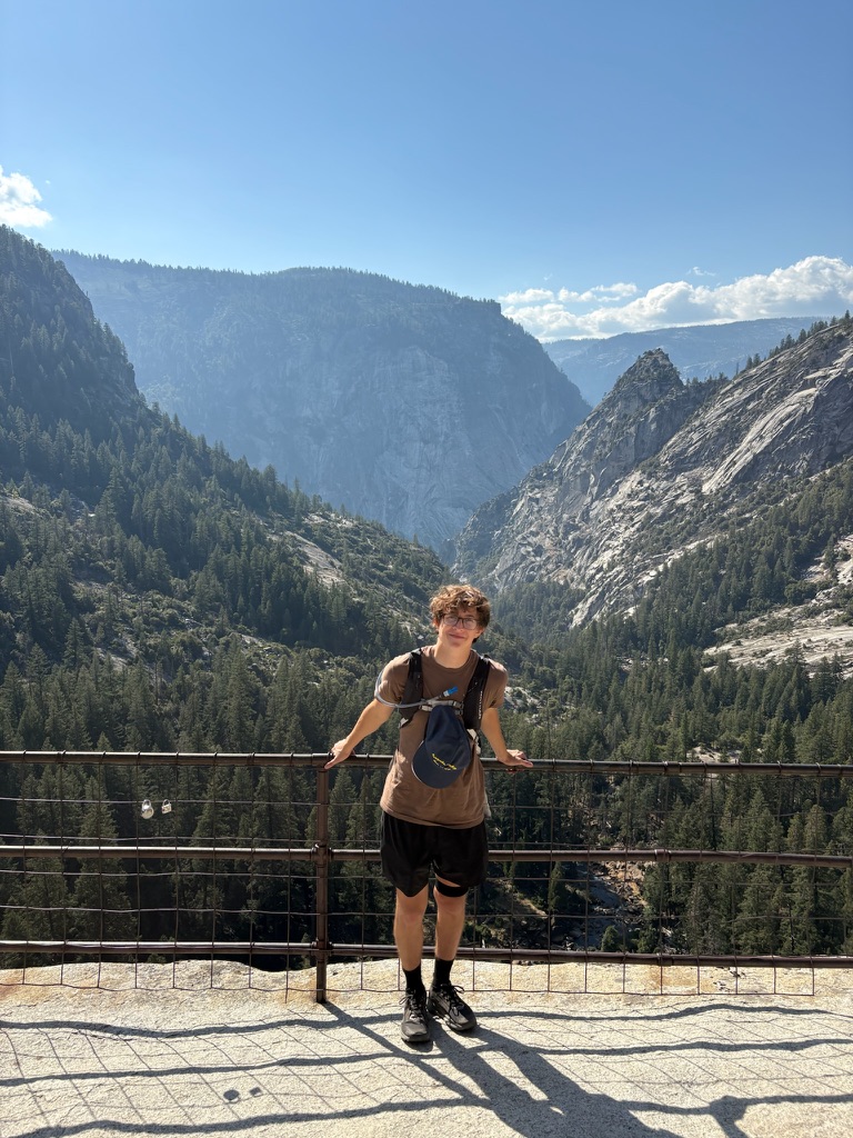 Young person with a backpack and cap hanging from chest standing by a railing overlooking a forested mountain valley under a clear blue sky.