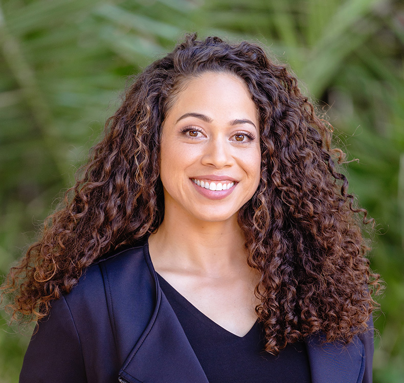 Smiling woman with long curly hair wearing a black top and jacket, standing outdoors with green foliage background.