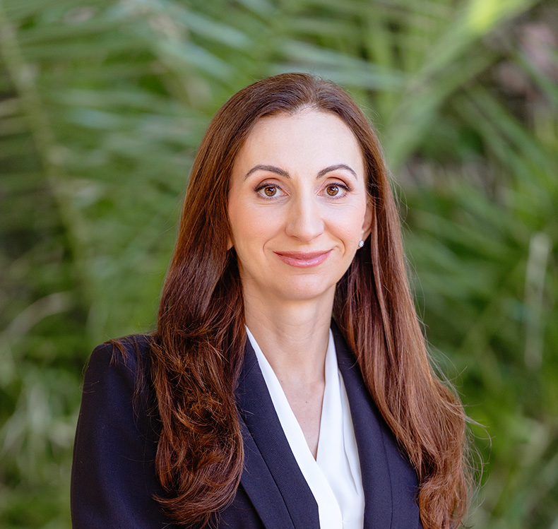 Professional woman with long brown hair wearing a dark blazer and white blouse standing in front of green foliage.