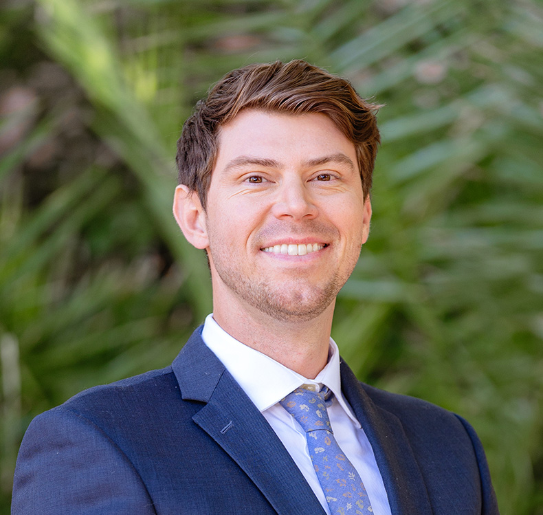 Smiling man in a blue suit and patterned tie standing outdoors with green foliage in the background.