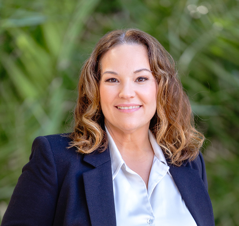 Smiling woman with curly light brown hair wearing a white blouse and dark blazer against a blurred green foliage background.