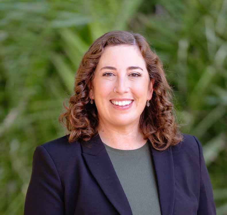 Woman with curly brown hair wearing a dark blazer and green top, posing outdoors with greenery background.