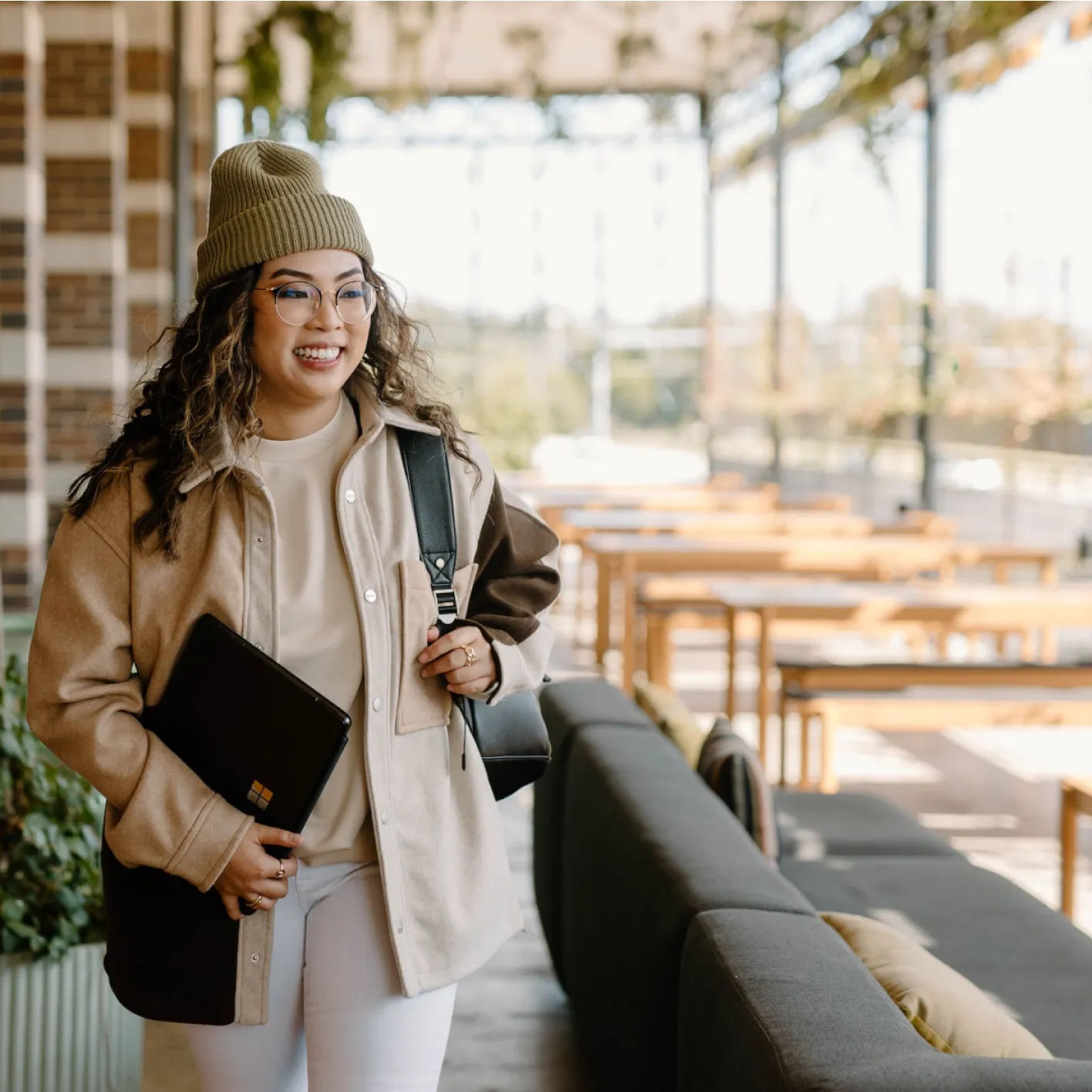 Jeune femme souriante avec un bonnet et des lunettes, tenant un ordinateur portable noir et portant un sac à dos dans un espace lumineux avec des tables en bois.