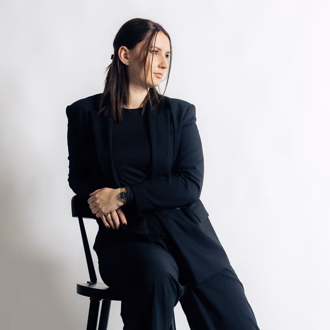 Zamišljena ženska z ravnimi rjavimi lasmi v črnem kostimu sedi na stolu in gleda vstran na belem ozadju.