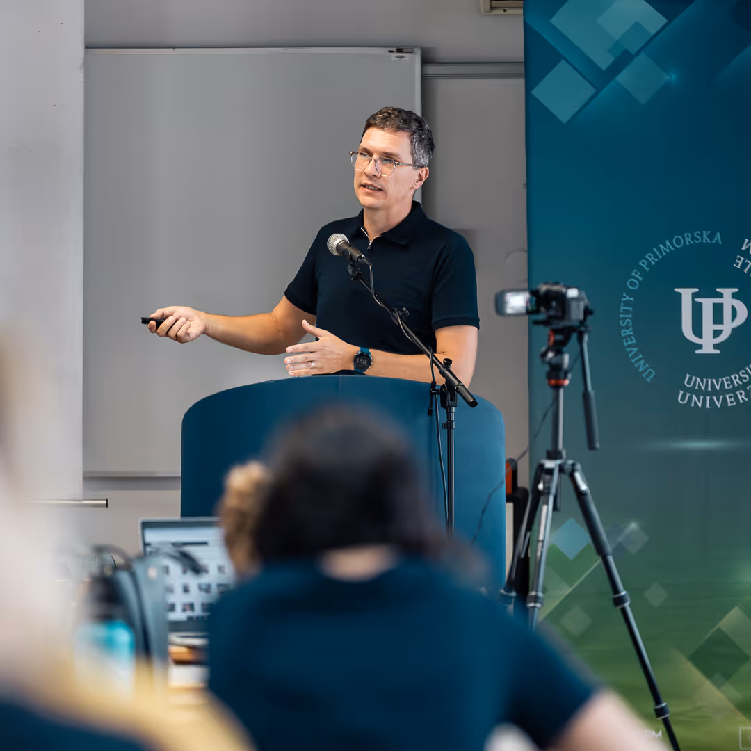 Moški z očali predava za govorniškim pultom z mikrofonom, medtem ko ga kamera na stojalu snema, v ozadju pa je transparent Univerza na Primorskem.