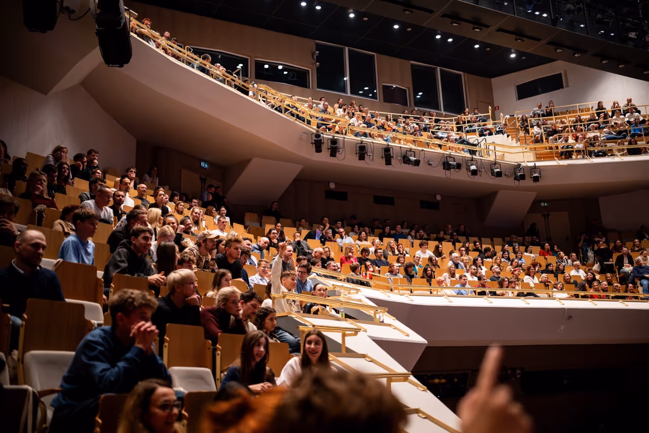 Polna večnivojska dvorana z ljudmi, ki sedijo in pozorno spremljajo dogodek.