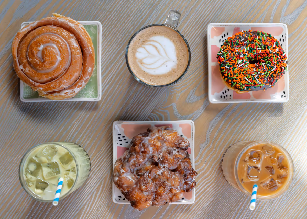 Assorted pastries and coffee drinks at The Hen cafe in Michigan, featuring a cinnamon roll, sprinkle donut, apple fritter, iced matcha, latte, and iced coffee on a wooden table.