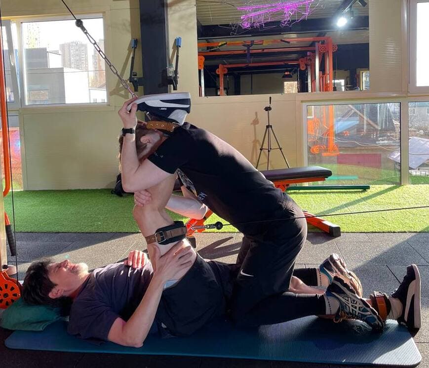 Індивідуальна робота в Dzhevaha Gym: контрольований стретчинг з підтримкою тренера для покращення рухливості кульшового суглоба та зменшення напруги у поперековому відділі. Позиція дозволяє безпечно збільшити амплітуду руху та відчути різницю між пасивним (статичним) розтягненням і активною (динамічною) роботою м’язів. Фото ілюструє підхід до теми: статичний vs динамічний стретчинг для спини при сидячій роботі. Статичні пози допомагають знизити тонус і «відпустити» перевантажені структури, тоді як динамічні рухи активують м’язи-стабілізатори та покращують контроль корпусу. Оптимальний результат досягається через грамотне поєднання обох методів залежно від стану спини та цілей клієнта.