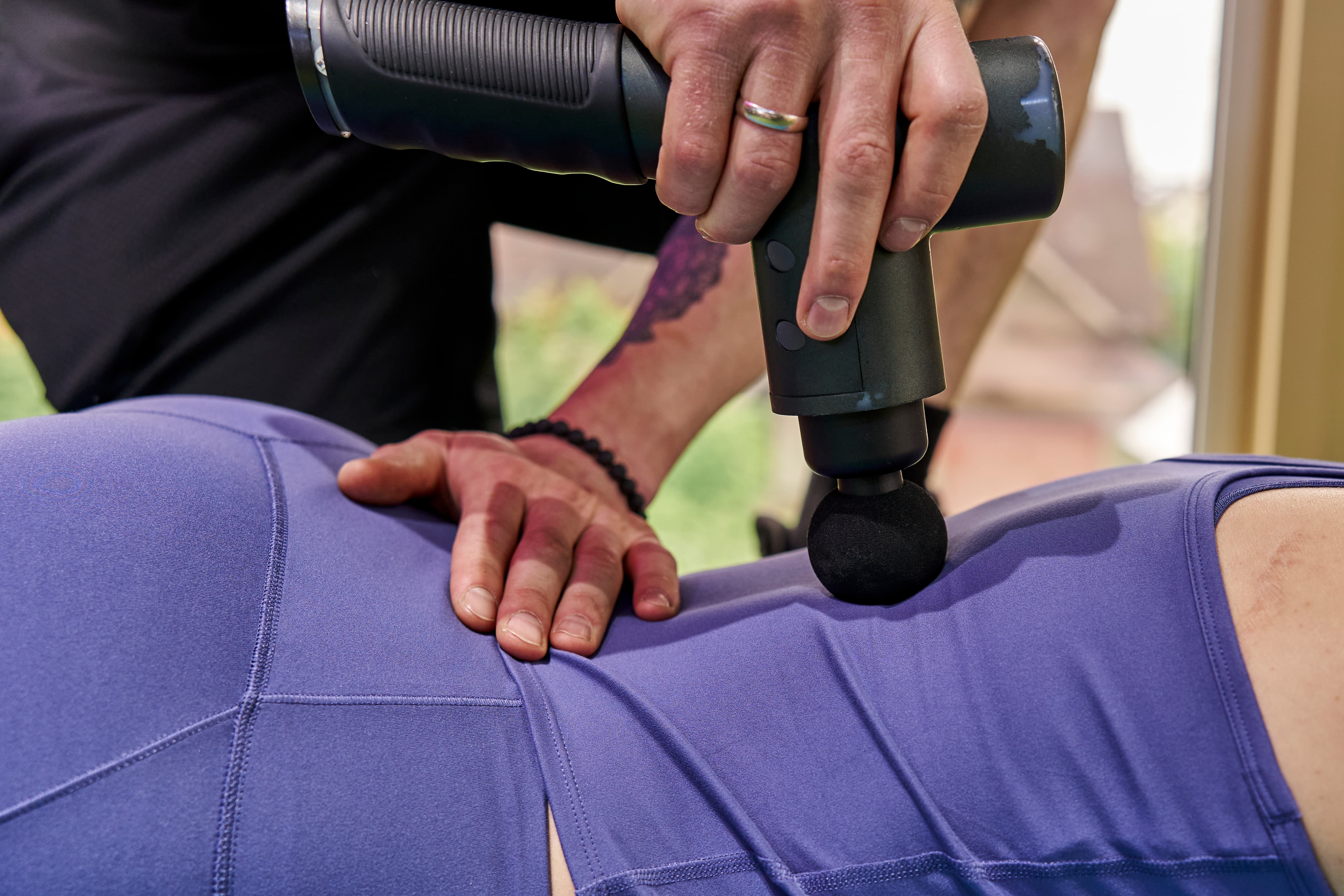 Крупний план: використання перкусійного масажера (massage gun) для міофасціального впливу на ділянку стегна/тазу. Насадка у формі кулі контактує з м’якими тканинами через одяг, створюючи ритмічний тиск. Рука спеціаліста стабілізує тканини поруч, що вказує на контрольовану, терапевтичну роботу з фасцією та м’язами.  Акцент: локальний вплив, робота з тригерними зонами, покращення ковзання тканин і зниження тонусу.  Ключові слова: перкусійна терапія, massage gun, міофасціальний реліз, фасція, тригерні точки, відновлення, кровообіг, зниження напруги, мобільність.  Безпека: не використовувати на кісткових виступах і суглобах, уникати надмірного тиску, тривалість впливу на одну ділянку 15–60 секунд, обережно при підвищеній чутливості або запаленні.