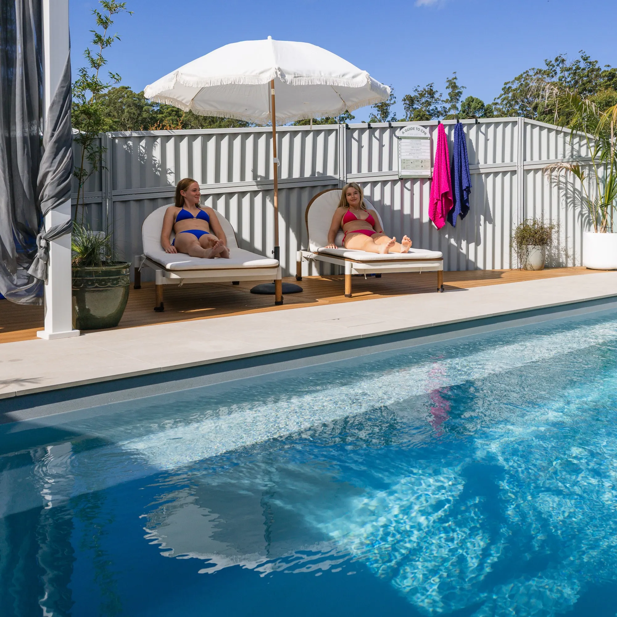 Two women in bikinis lounging on white pool chairs under a white umbrella beside a clear blue swimming pool.
