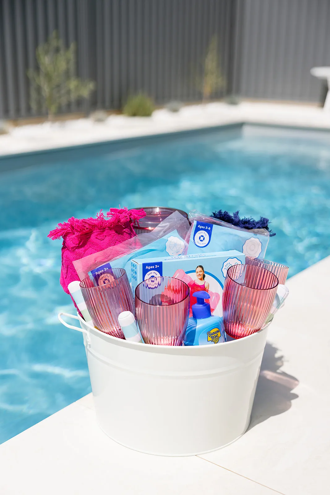 White bucket filled with pink glasses, sunscreen, pool toys, and a pink towel placed by a swimming pool.