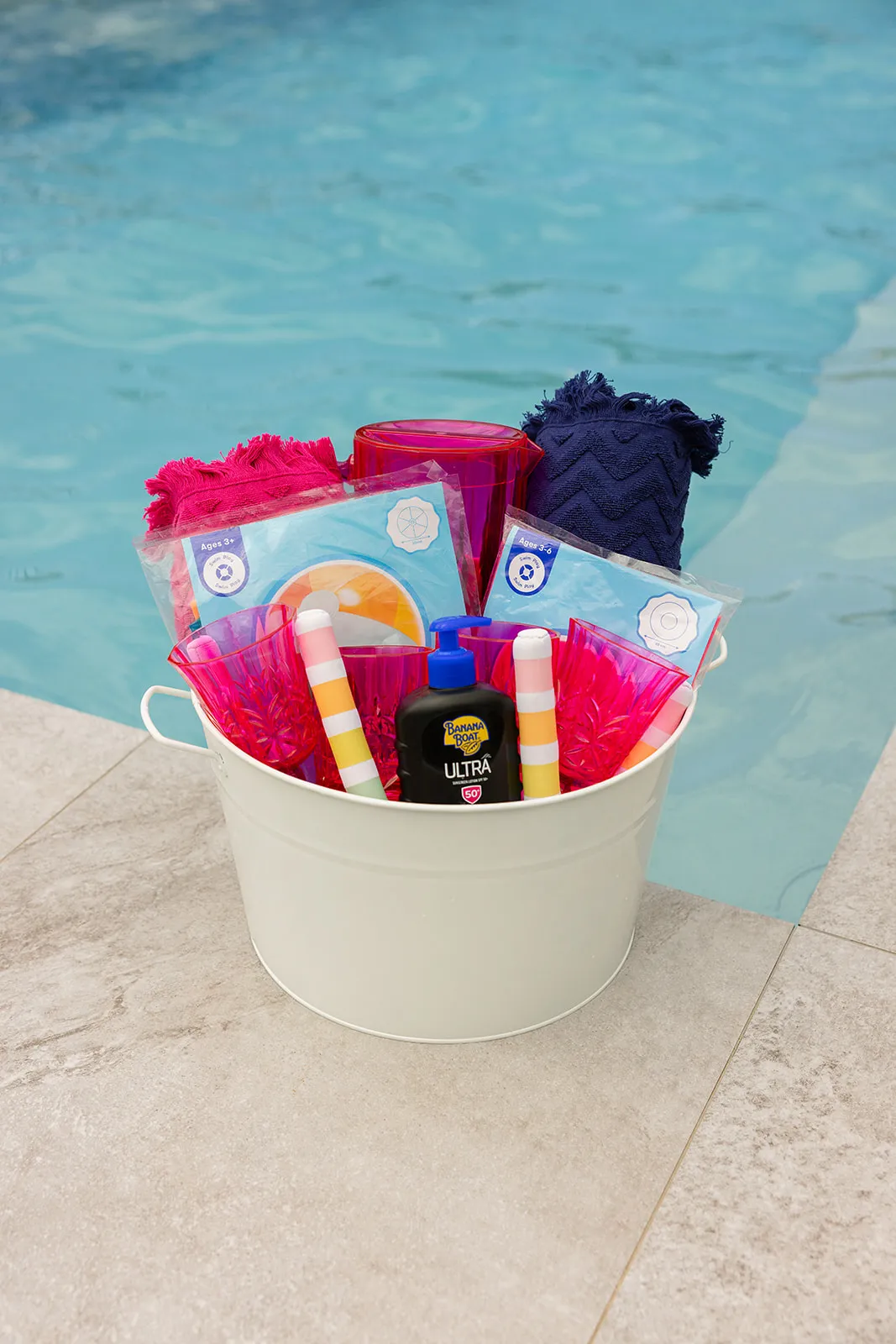 White bucket by pool edge containing rolled towels, pink plastic cups, sunscreen, and packaged pool toys.