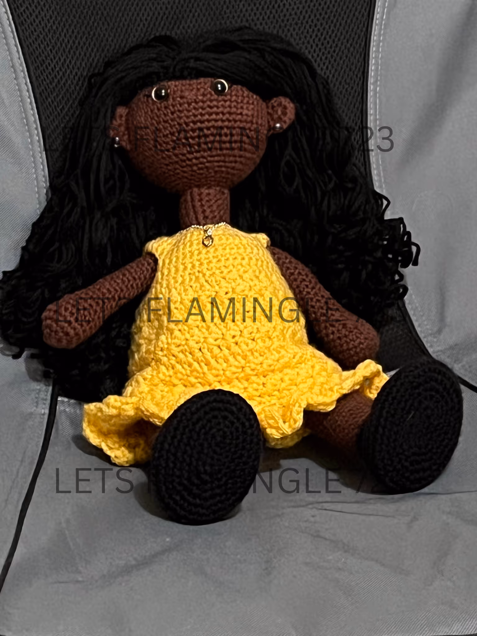 Handmade crocheted doll with dark brown skin, long black yarn hair, wearing a yellow dress and black shoes seated on a gray fabric surface.