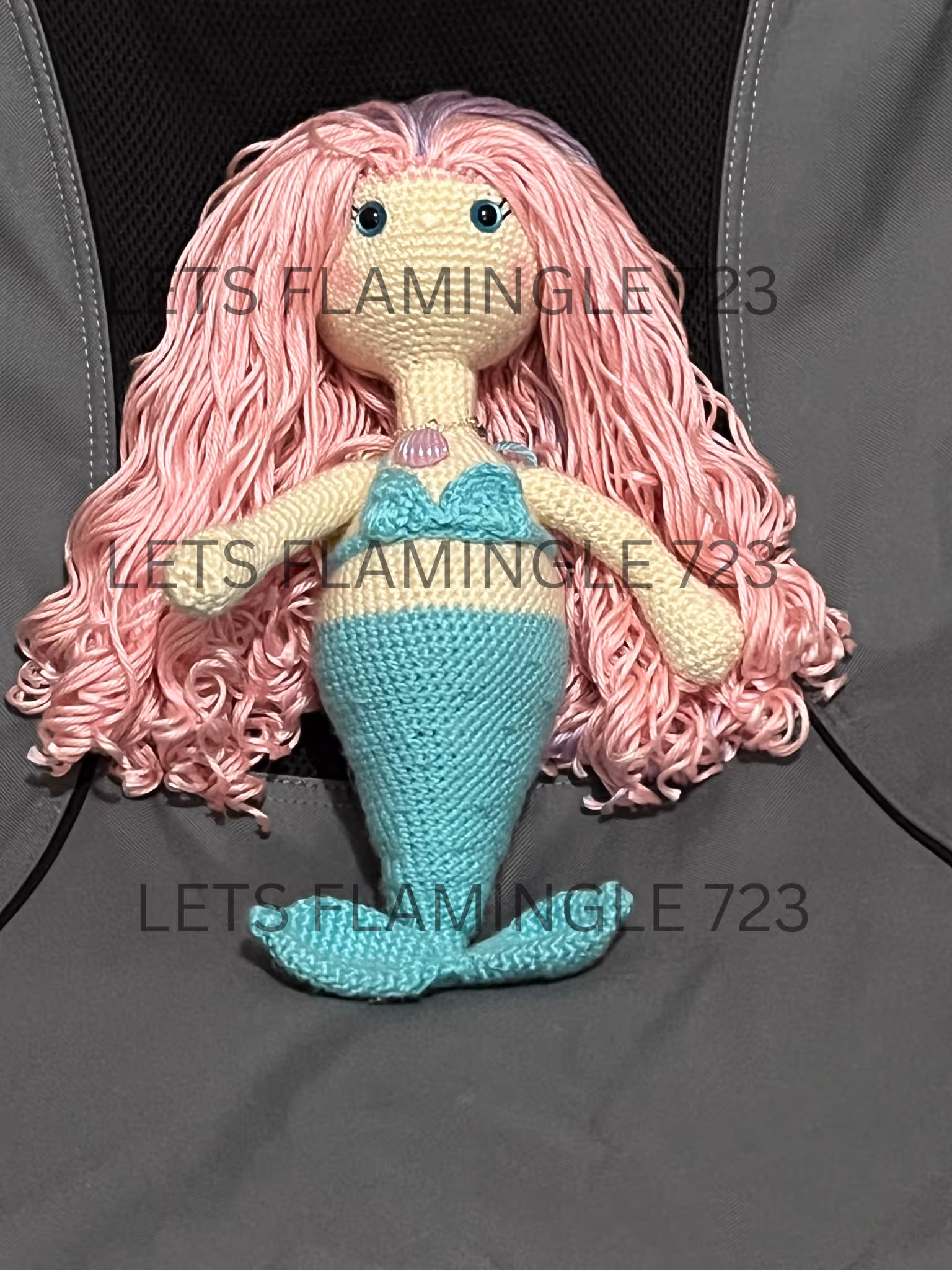 Hand-crocheted mermaid doll with long pink curly hair, teal tail, and matching bikini top, sitting on a gray surface.