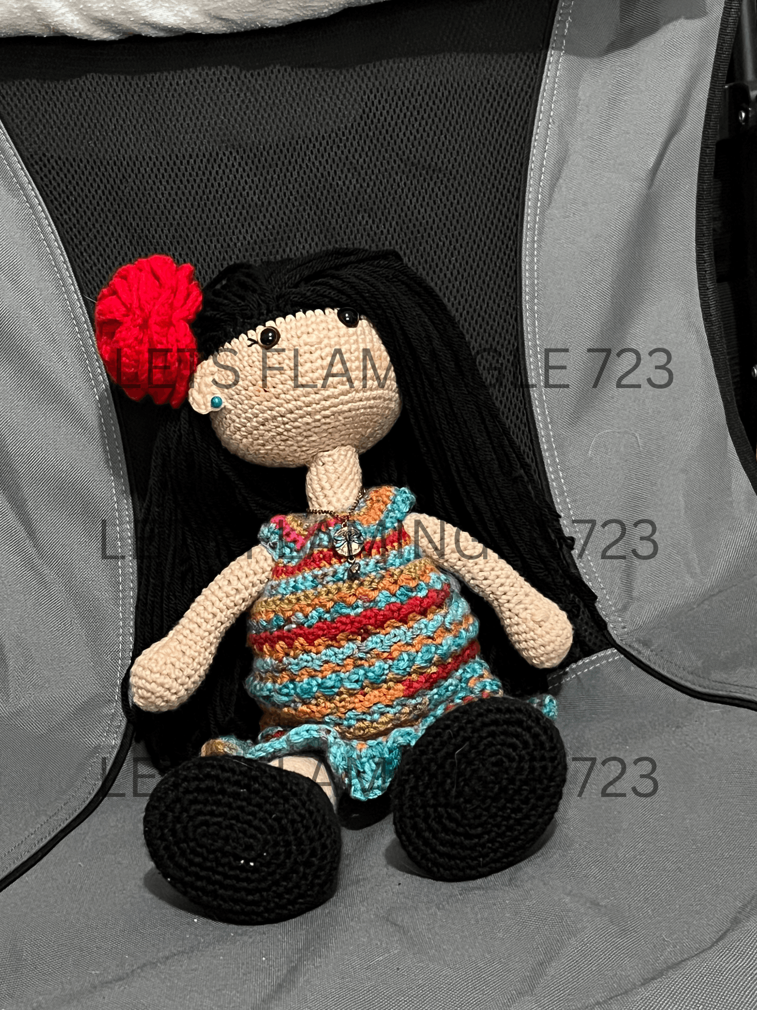 Crocheted doll with long black hair, a multicolored striped dress, black shoes, and a red flower in her hair sitting on a gray fabric chair.