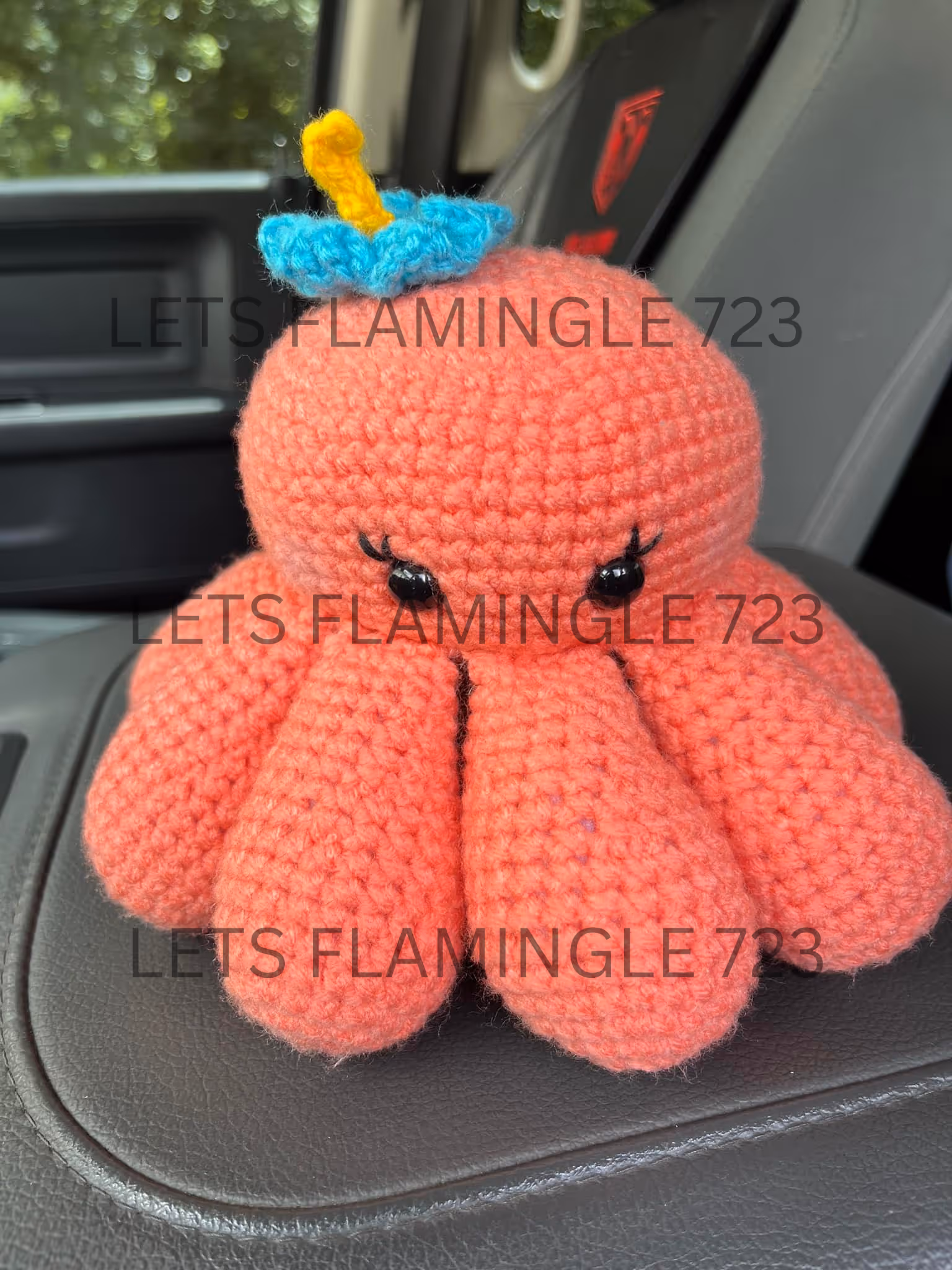 Crocheted pink octopus plush with black eyes and a blue and yellow flower on its head sitting on a car armrest.