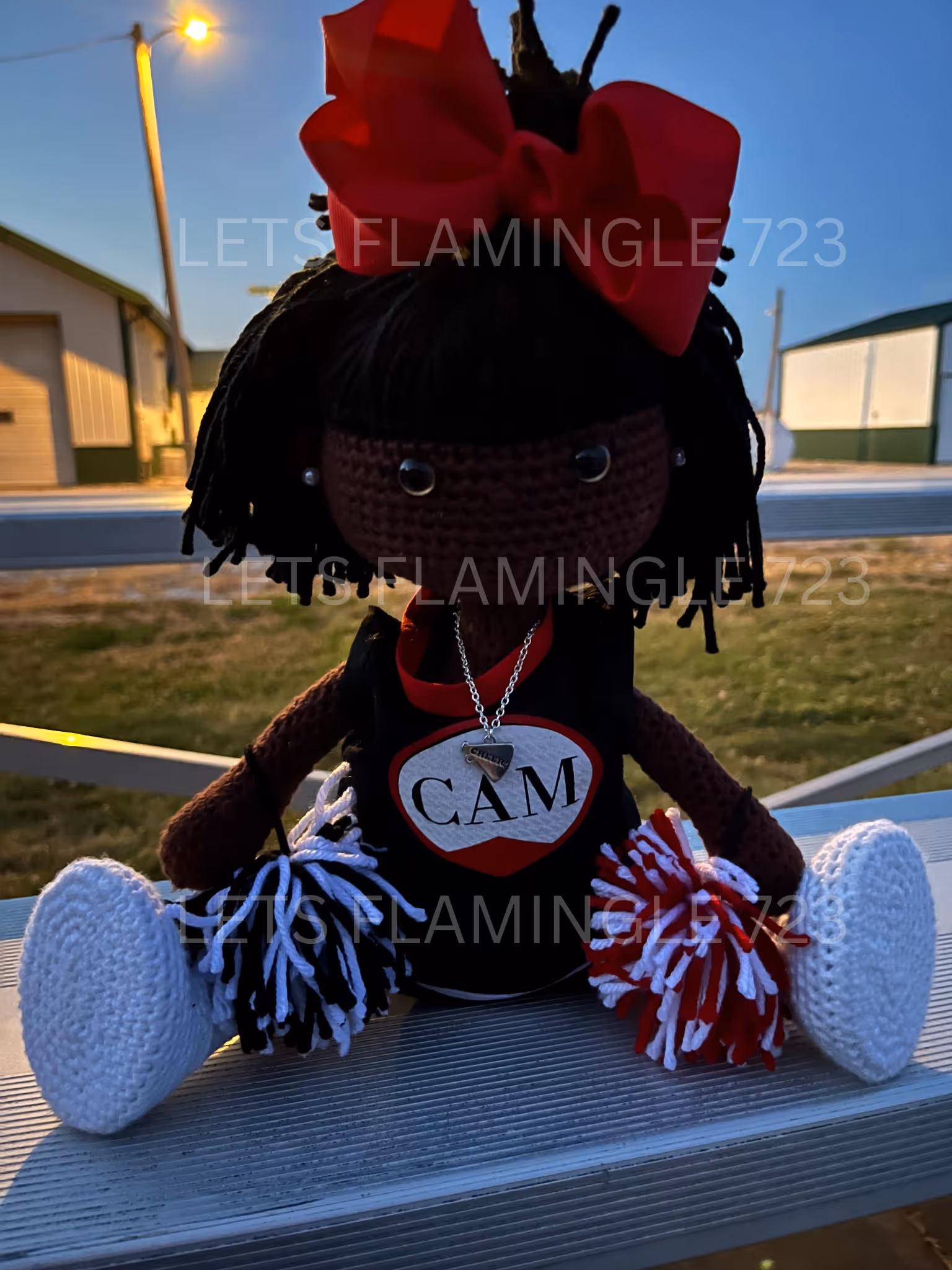Handmade crocheted cheerleader doll with black yarn hair, a large red bow, white shoes, and red and white pom-poms sitting on metal bleachers.