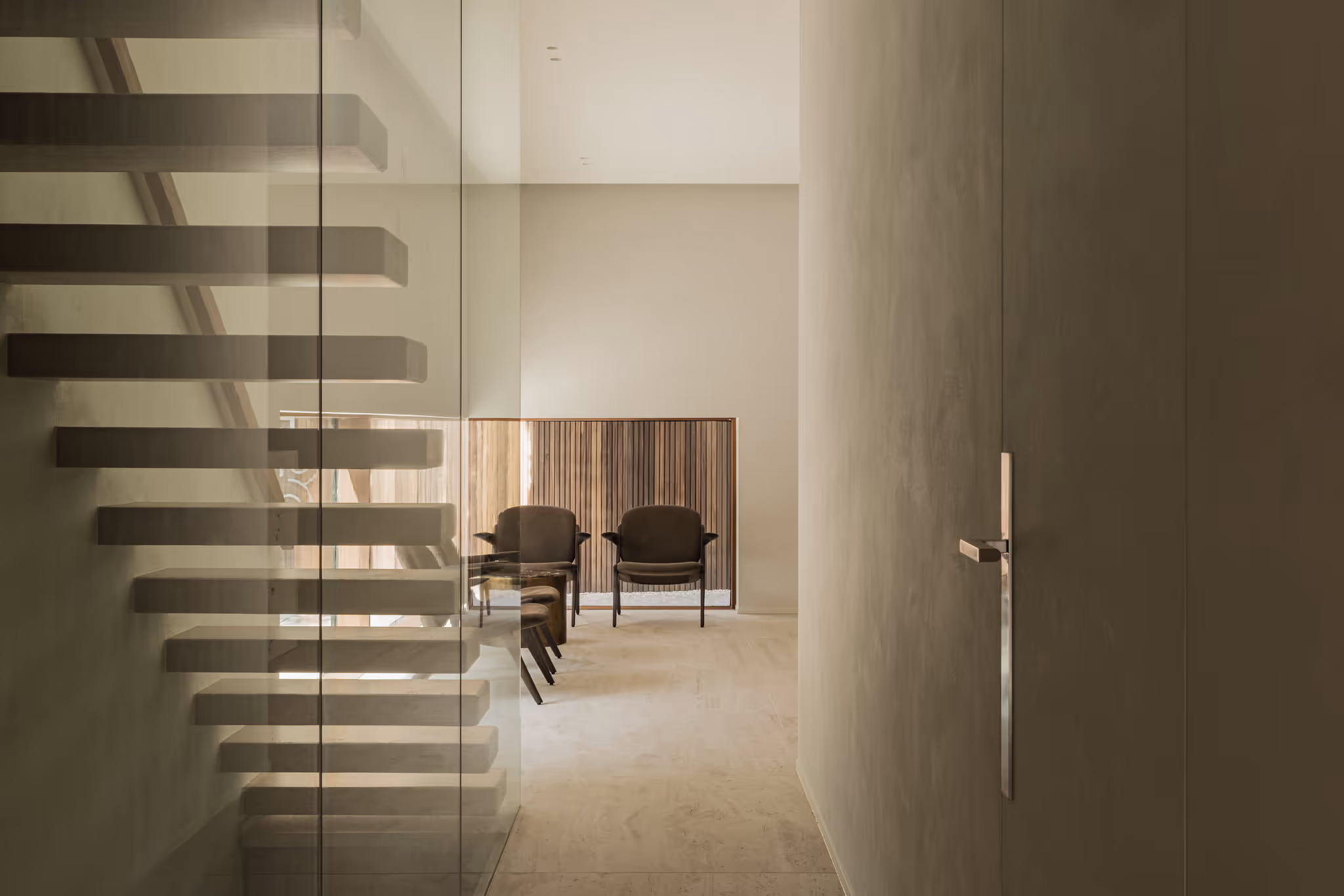 Minimalist hallway with floating staircase behind a glass wall. At the end of the hallway are two modern chairs by a wooden slatted wall.