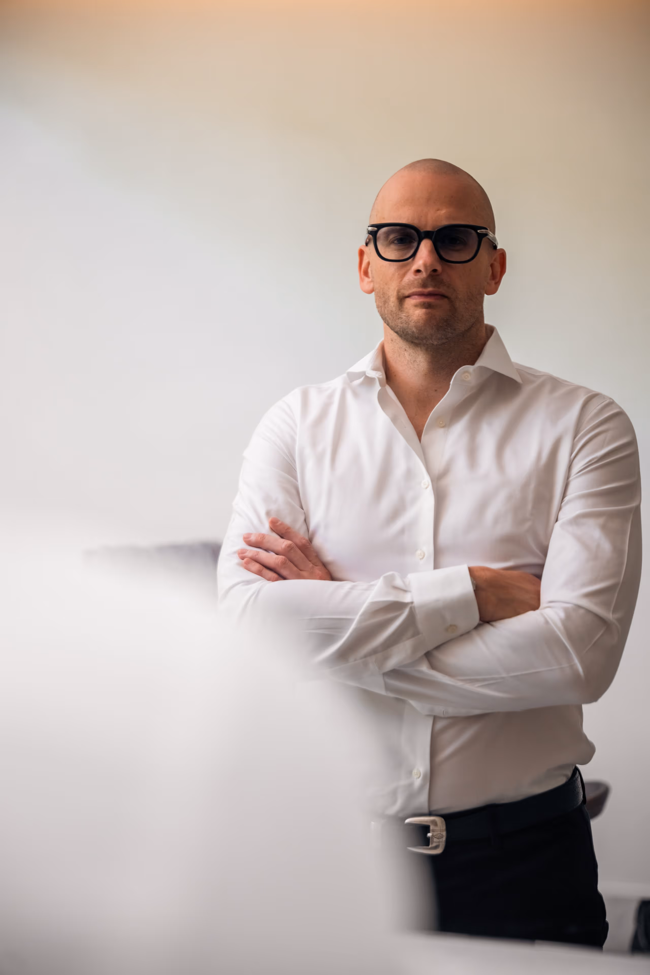 Portret van Dr. Maarten Van Genechten met een witte blouse en een bril, staand met armen over elkaar en een neutrale blik.