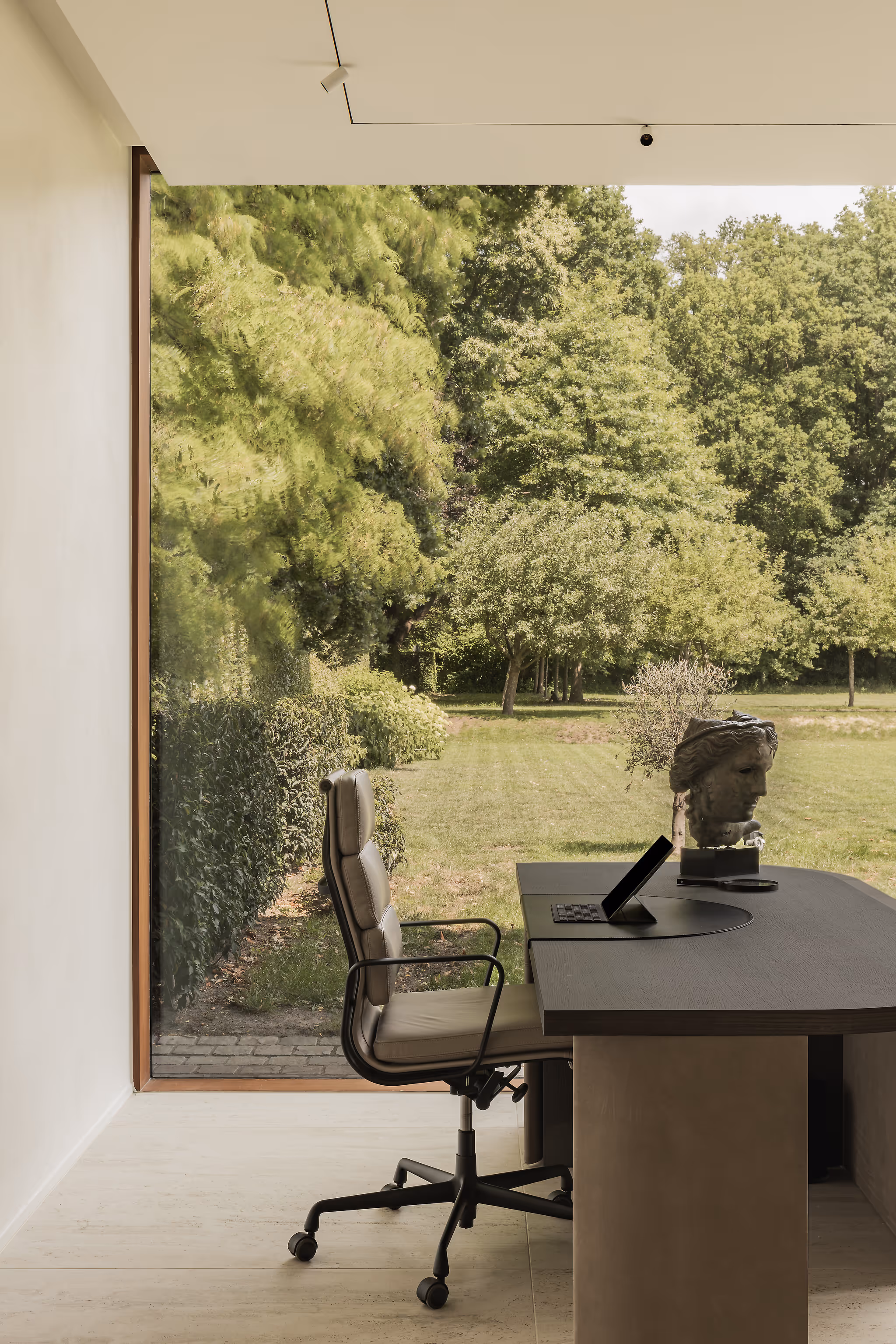 Modern office overlooking a green garden through a large glass wall. At the desk is an ergonomic office chair and on the desk is a classic sculpture.