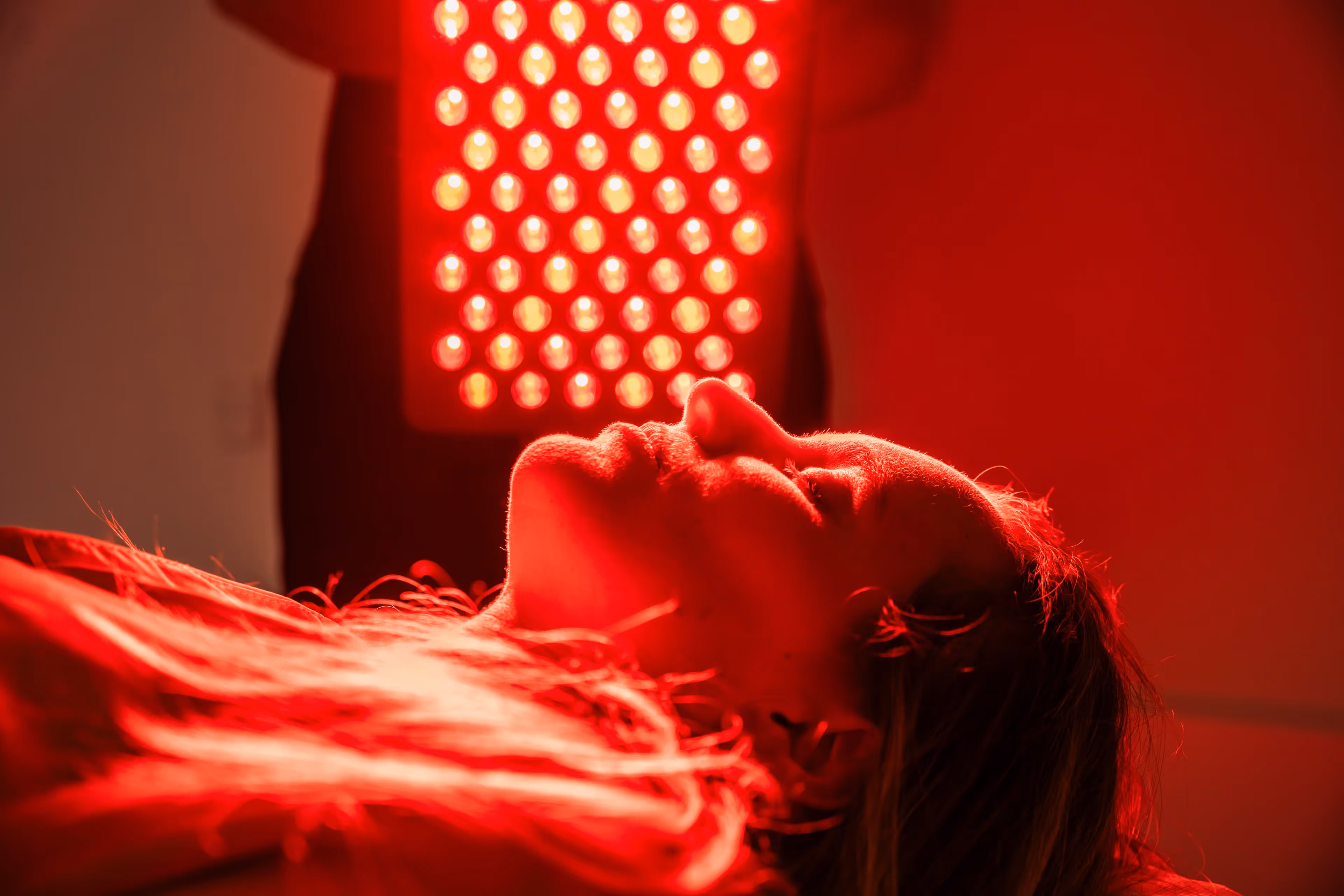 Close-up van een vrouw die onder een fel rood licht ligt tijdens een lichttherapiebehandeling. Het rode LED-paneel is prominent in beeld.