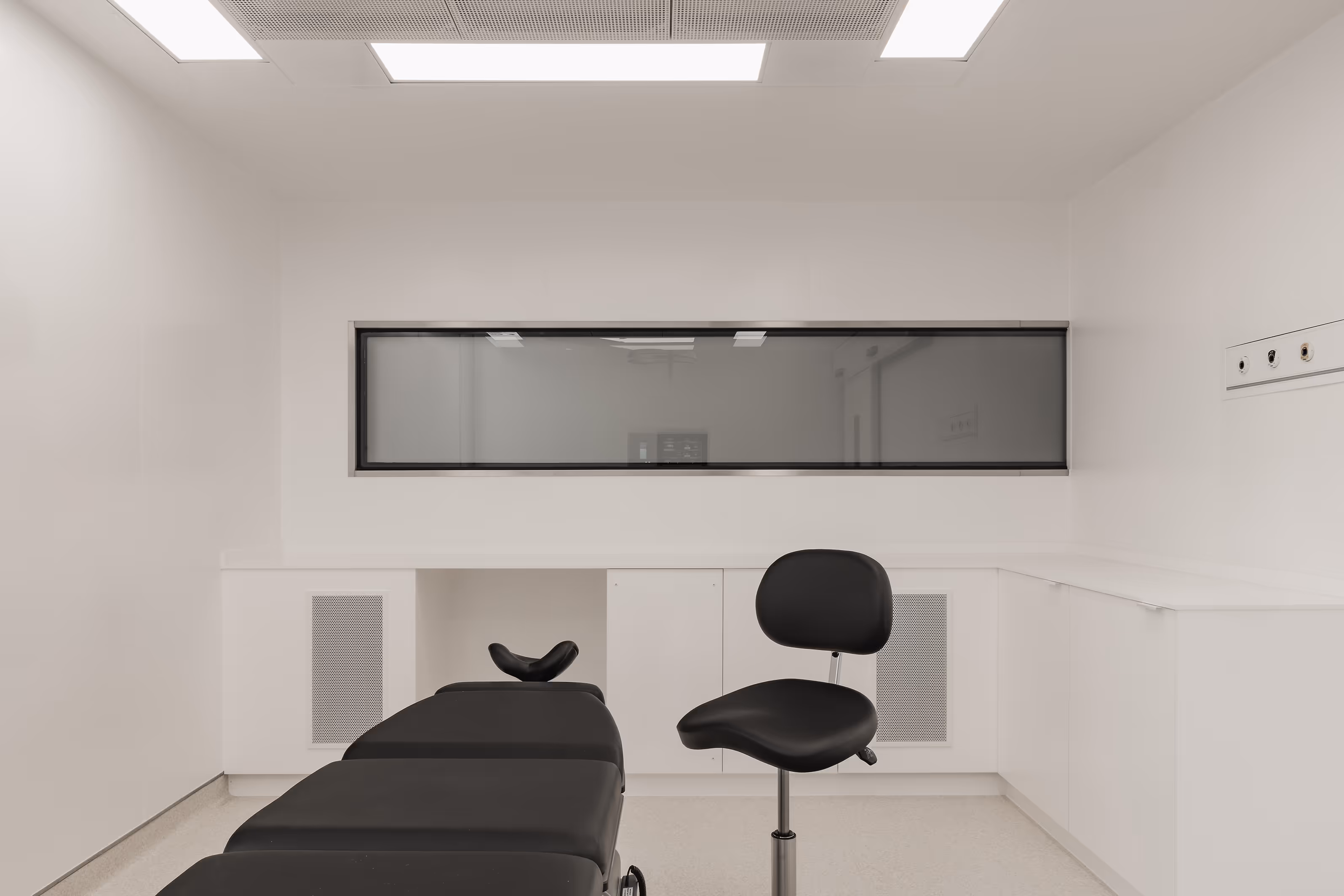 Een behandelruimte met een zwarte behandeltafel en zwarte kruk. Aan de muur een groot raam dat uitzicht biedt naar een andere ruimte.