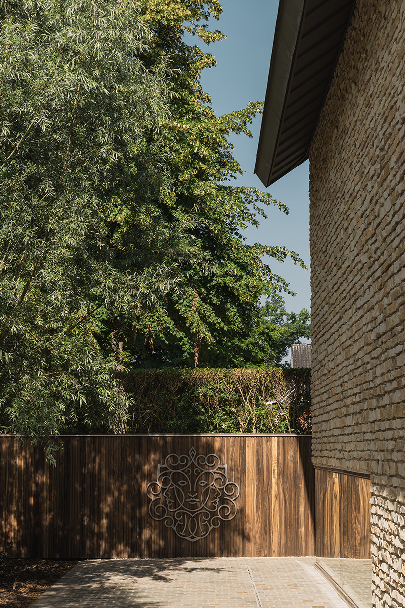 Een moderne houten tuinpoort met een decoratief metalen embleem, omgeven door groene bomen en een haag. Rechts is een muur van natuursteen zichtbaar.