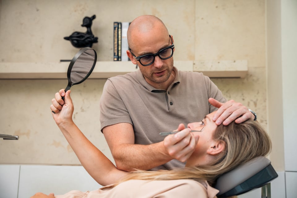Le Dr Maarten Van Genechten traite le visage d'une femme allongée sur une chaise de traitement. La femme brandit un miroir à main rond pour voir son visage.