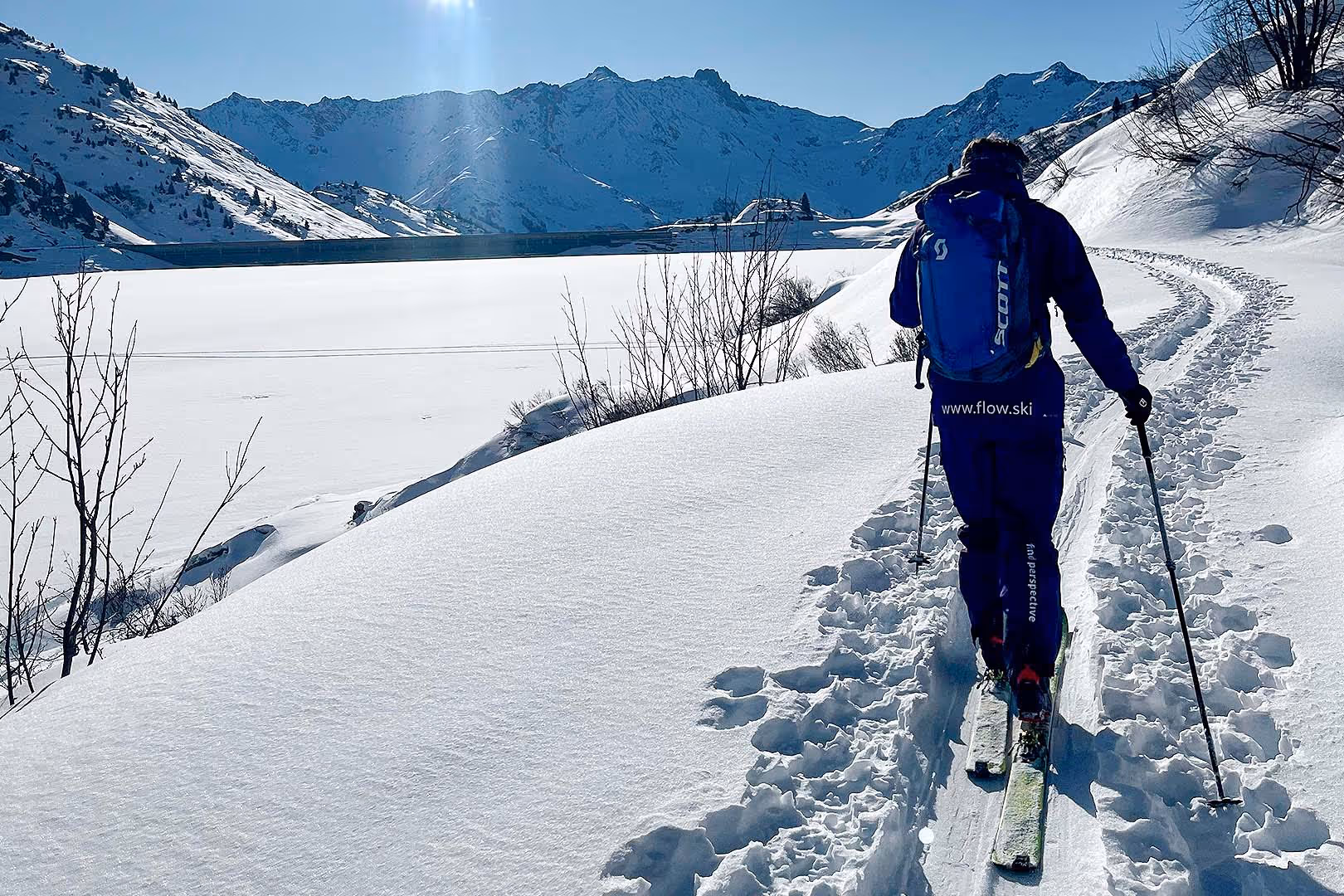 Skilehrer von Flow in lila Kleidung mit Rucksack steigt auf verschneitem Pfad in den Bergen bei sonnigem Wetter auf.