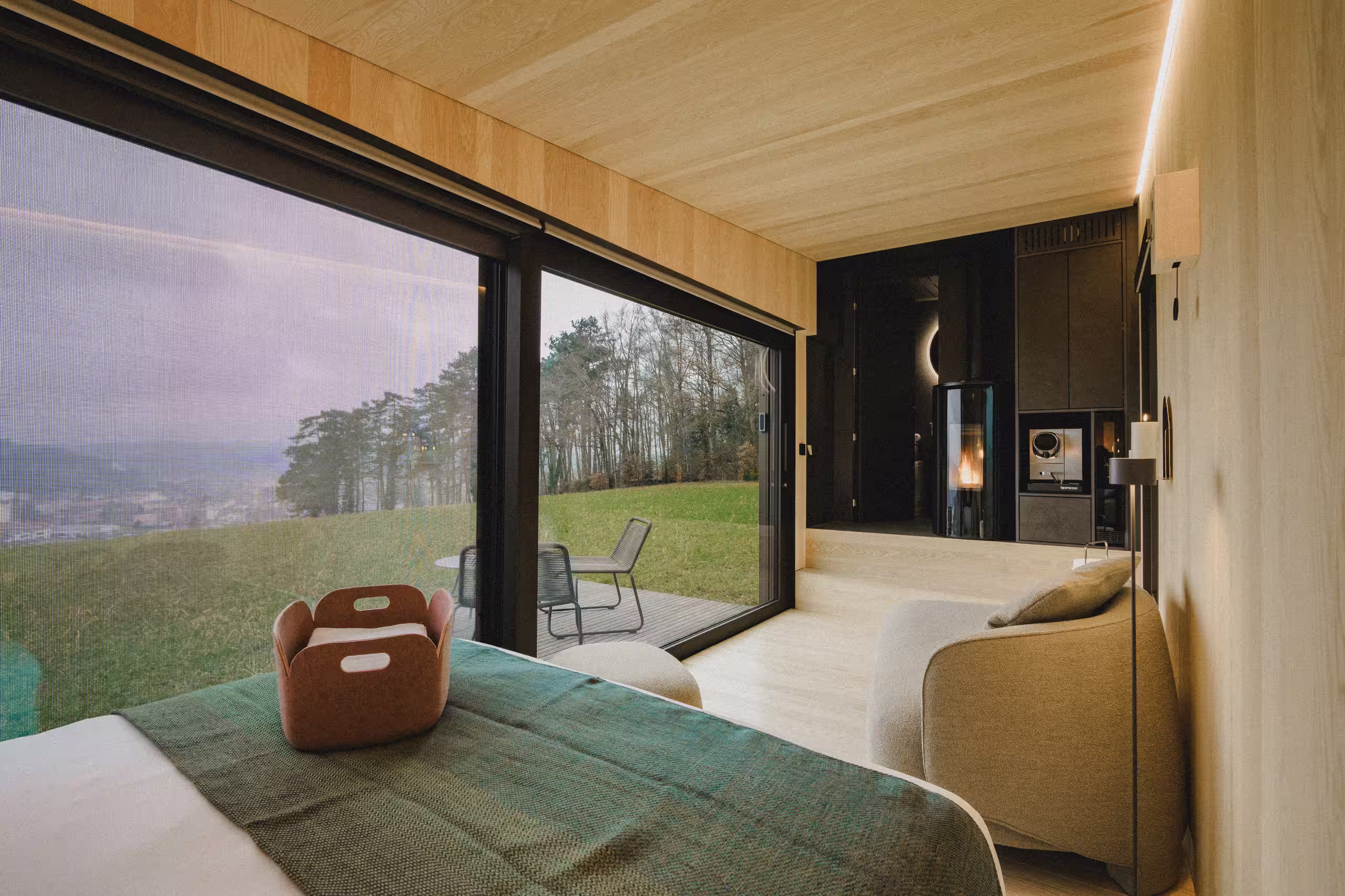 Interni accoglienti di una cabina SNUUG con stufa a legna e grande vetrata che si apre sulla foresta svizzera, che offre il comfort di un hotel a 4 stelle.