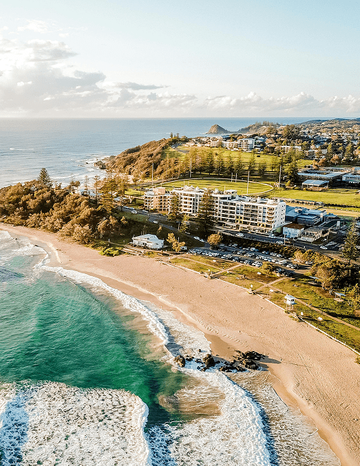 Aerial view of a coastal town with sandy beach, turquoise ocean waves, residential buildings, and green hills under a partly cloudy sky.